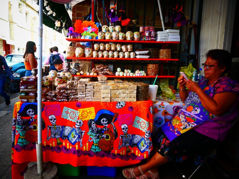 A woman sits beside a Dia de los Muertos stall. The table has a red tablecloth embroidered with skeletons, and on shelves hanging above, there are sugar skulls on sale to eat.