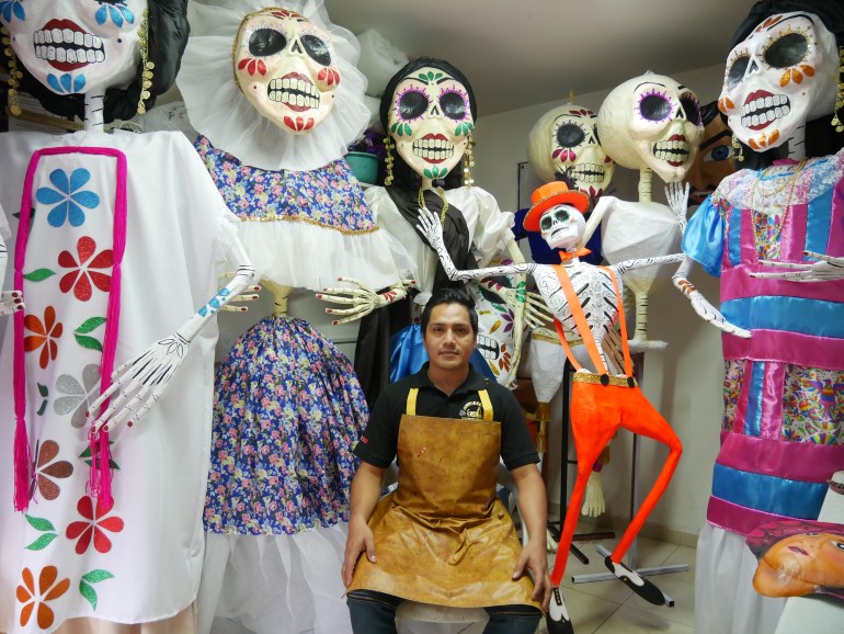 A man in a brown apron sits in a room crowded with paper-mache giants, depicting large skeletal figures like La Calavera Catrina.